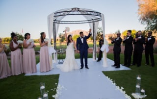Newlywed Bride and Groom conclude ceremony by raising their arms and greeting guests as husband and wife. Bridesmaids and Groomsmen clap and cheer on either side of the altar as the sun sets.