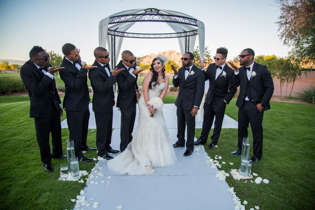 Bride and Groomsmen take a funny portrait photo. The Groomsmen wear sunglasses as they blow kisses at Bride while she poses sweetly, surrounded by ivory rose petals. Las Vegas wedding photography.