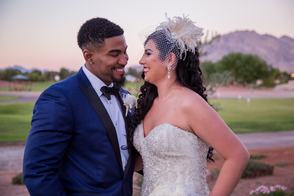 Fashion photographer captures a joyful moment between Bride and Groom at sunset. The Groom is wearing a handsome blue and black color block tuxedo jacket. The Bride is wearing a lovely vintage birdcage visor and drop pearl earrings.