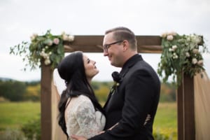 Rustic wedding photography. Bride and Groom embrace each other beneath a florally decorated atrium in an open field of wildflowers on an overcast day.