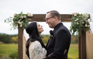 Rustic wedding photography. Bride and Groom embrace each other beneath a florally decorated atrium in an open field of wildflowers on an overcast day.