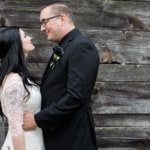 eivans_testimonial_100717__0141 Bride and Groom look loving at one another posed in front of a rustic wooden cabin. The Groom is wearing a black rose boutonniere.