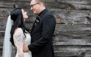 Bride and Groom look loving at one another posed in front of a rustic wooden cabin. The Groom is wearing a black rose boutonniere.