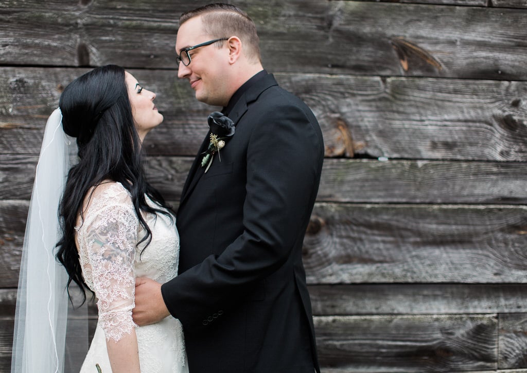 Bride and Groom look loving at one another posed in front of a rustic wooden cabin. The Groom is wearing a black rose boutonniere.