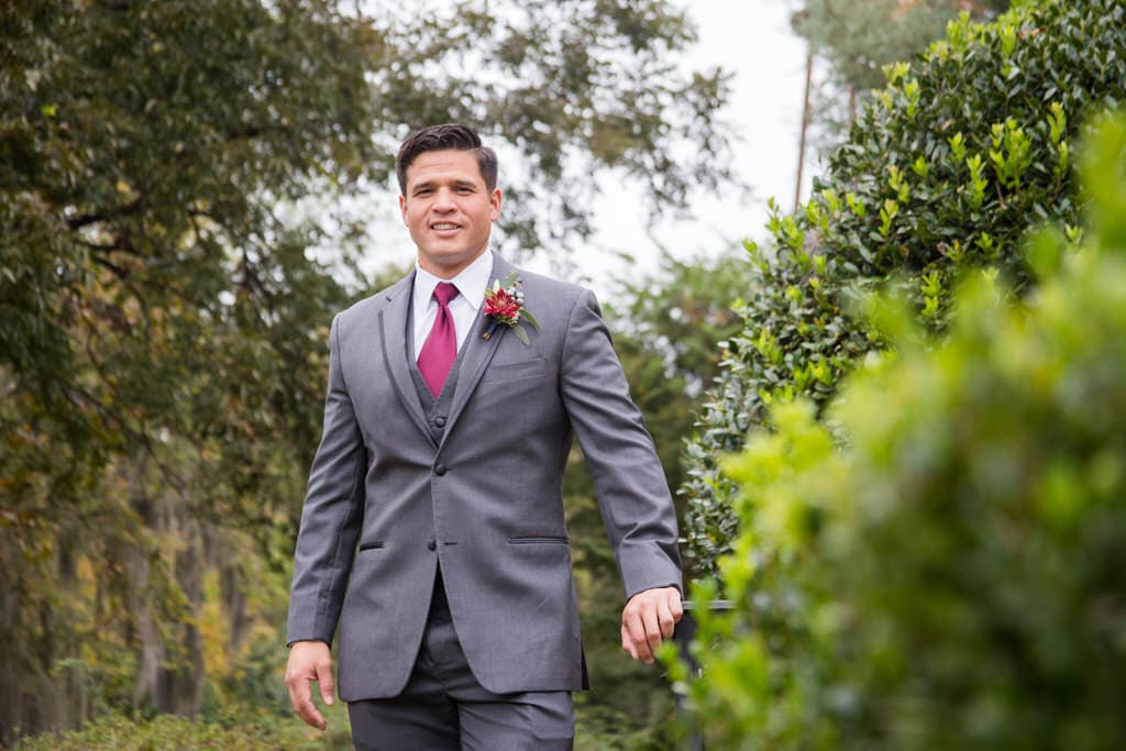 A handsome Groom sports a burgundy tie with a gray, three-piece suit and a smile. Trees and bushes cover the background as he prepares to marry.