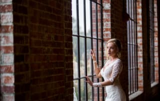 Fashion photographer captures a stunning shot of a Bride gazing out of a floor to ceiling window of an exposed brick loft. The bride has deep red lips and wears her hair in a loose and wavy updo with escaping tendrils.