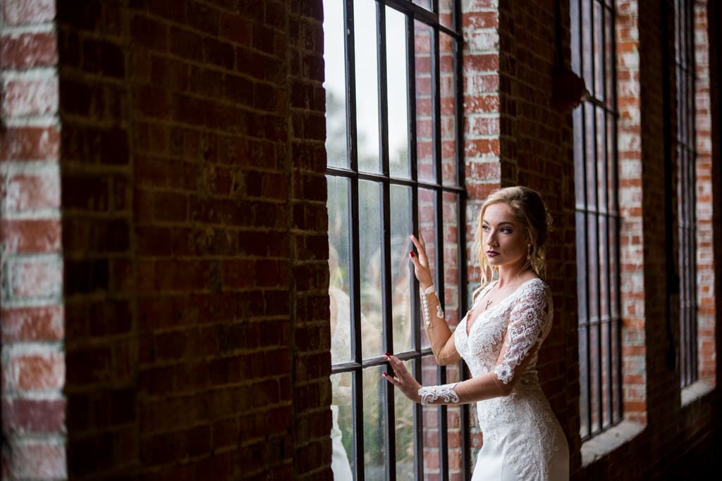 Fashion photographer captures a stunning shot of a Bride gazing out of a floor to ceiling window of an exposed brick loft. The bride has deep red lips and wears her hair in a loose and wavy updo with escaping tendrils.