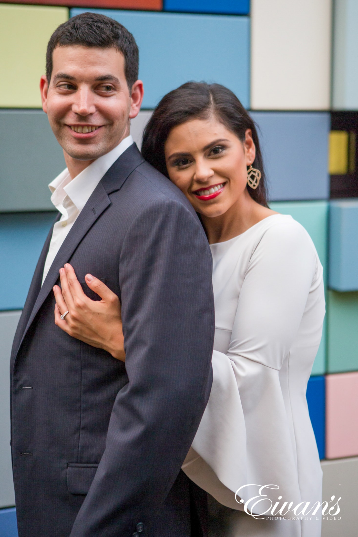 image of engaged man and woman, woman is hugging man from behind in front of a backdrop of colored squares