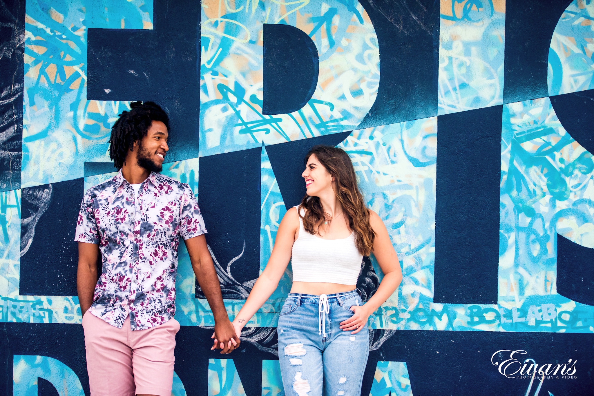 Beach Engagement Photos (223) woman in white and pink floral shirt and blue denim jeans standing beside wall with graffiti
